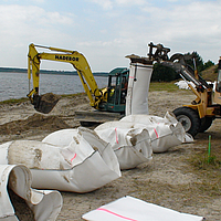 Riempimento con sabbia disponibile in loco dei sacchi SoilTain con l'aiuto di un escavatore mentre la pala gommata sostiene il sacco di sabbia prefabbricato