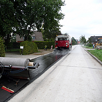 Una macchina a termine avanza, seguita dall'applicazione dell'armatura in asfalto SamiGrid® sulla strada
